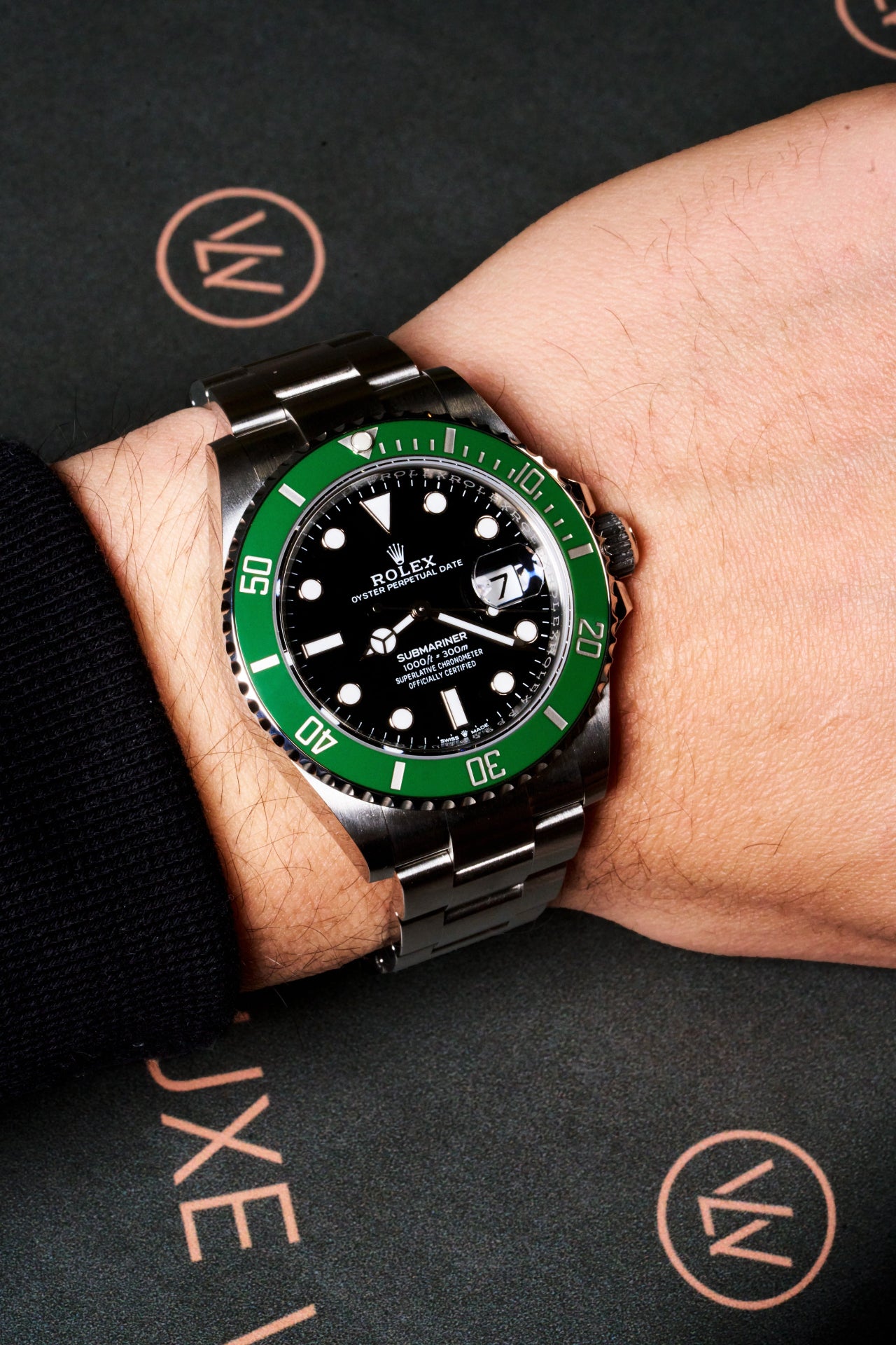 Close-up of a green-bezel Rolex Submariner watch on wrist, positioned over a dark green Luxewrist surface with copper-toned branding