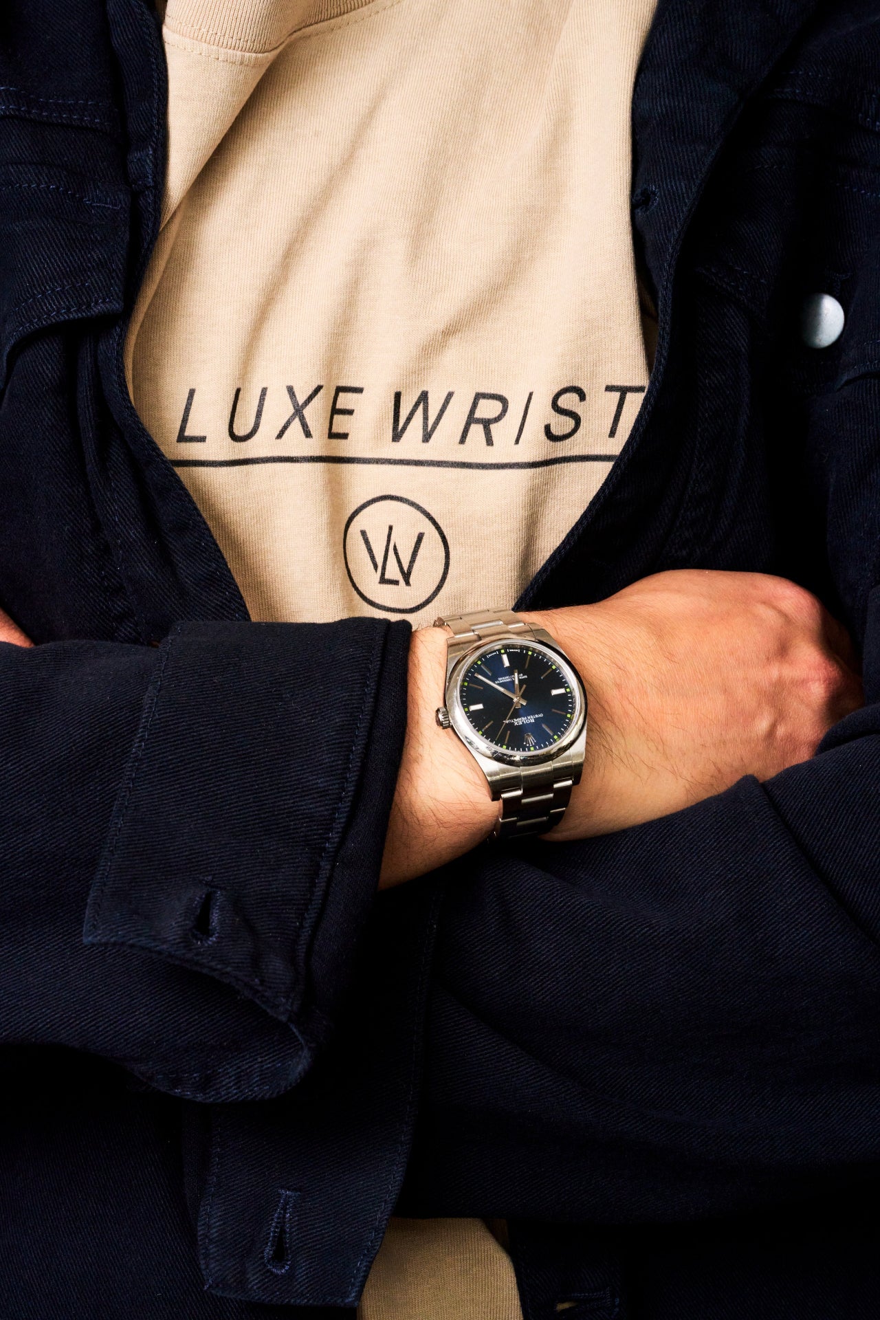 Close-up of a person wearing a tan Luxewrist logo tee layered under a dark jacket, paired with a classic stainless steel wristwatch with a black dial