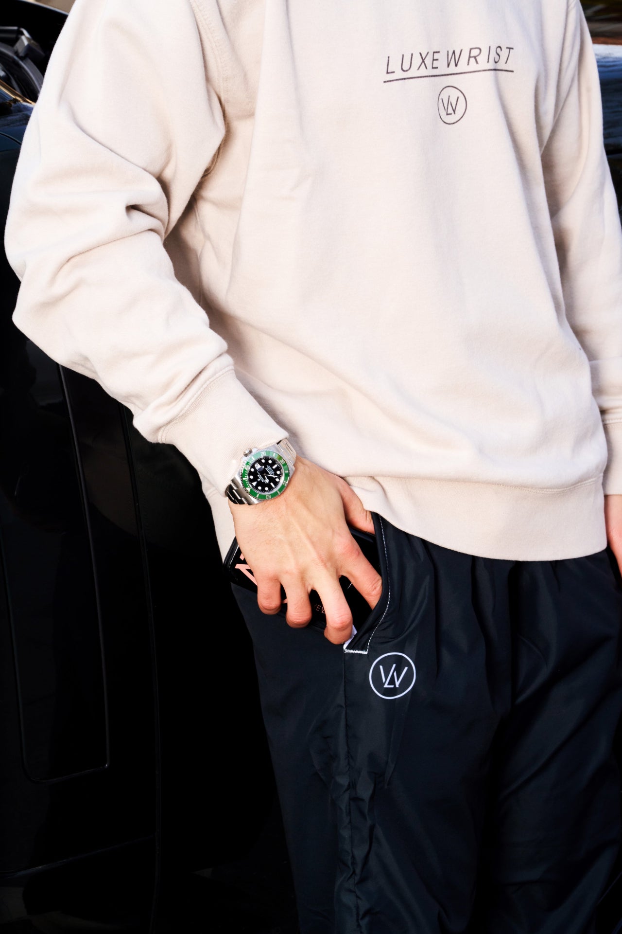 Man wearing a beige Luxewrist sweatshirt and navy trackpants with logo detail, styled with a bold green-faced luxury watch in an urban streetwear setting