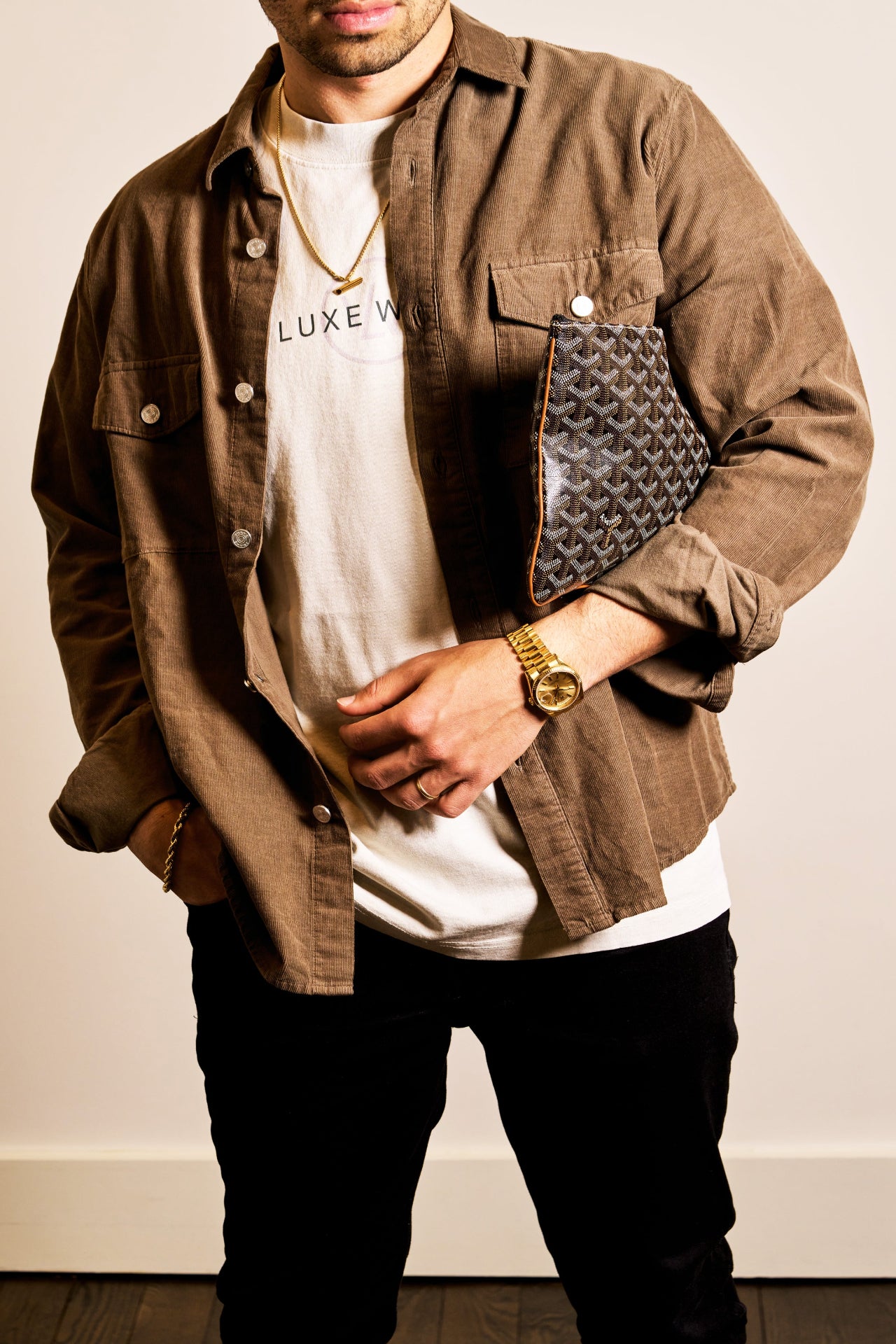 Stylish man wearing a Luxewrist logo tee layered under a brown corduroy overshirt, accessorized with a gold watch, gold chain, and a designer clutch bag