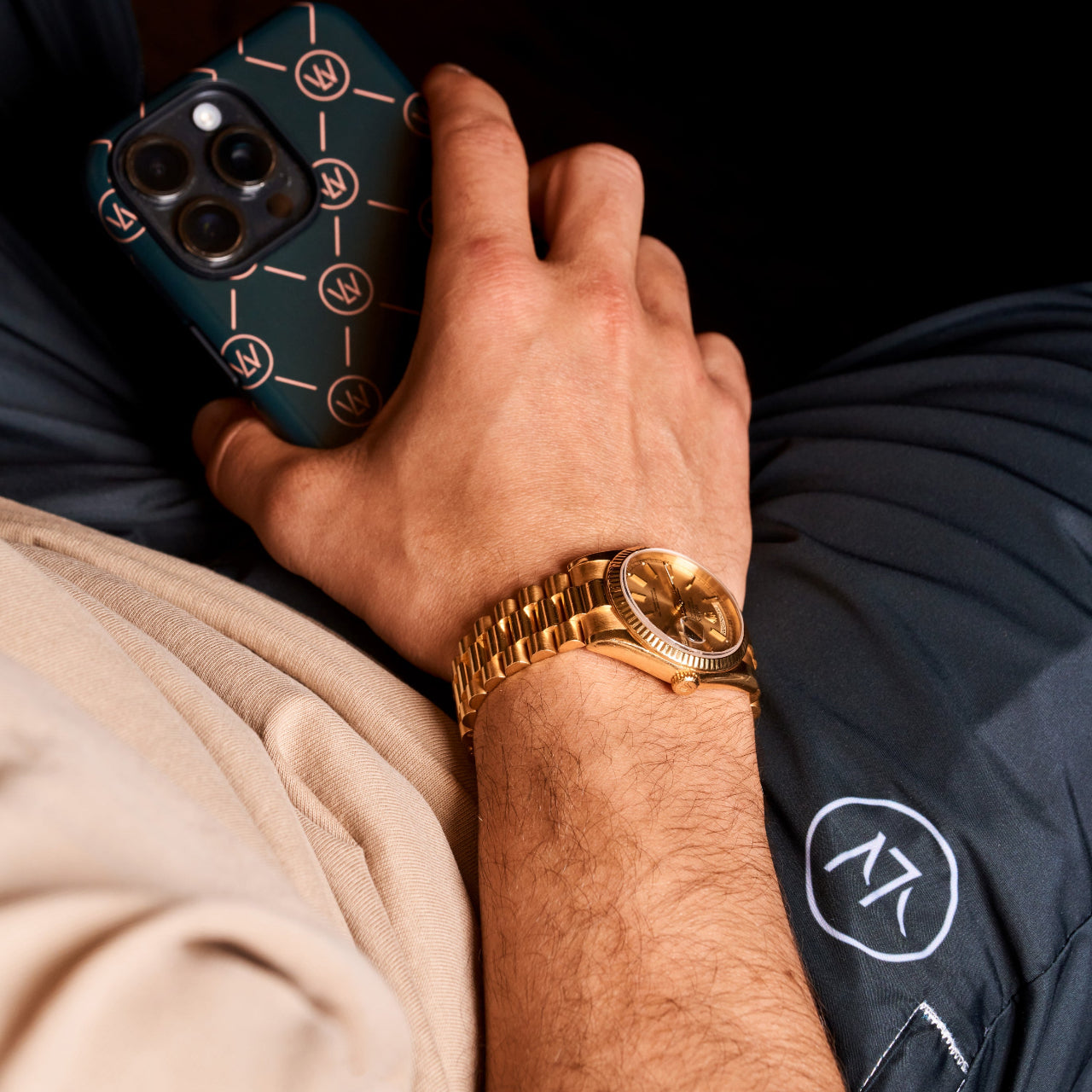 Relaxed lifestyle shot of a person wearing a gold wristwatch and holding a Luxewrist phone case, with branded navy trackpants and beige sweatshirt visible