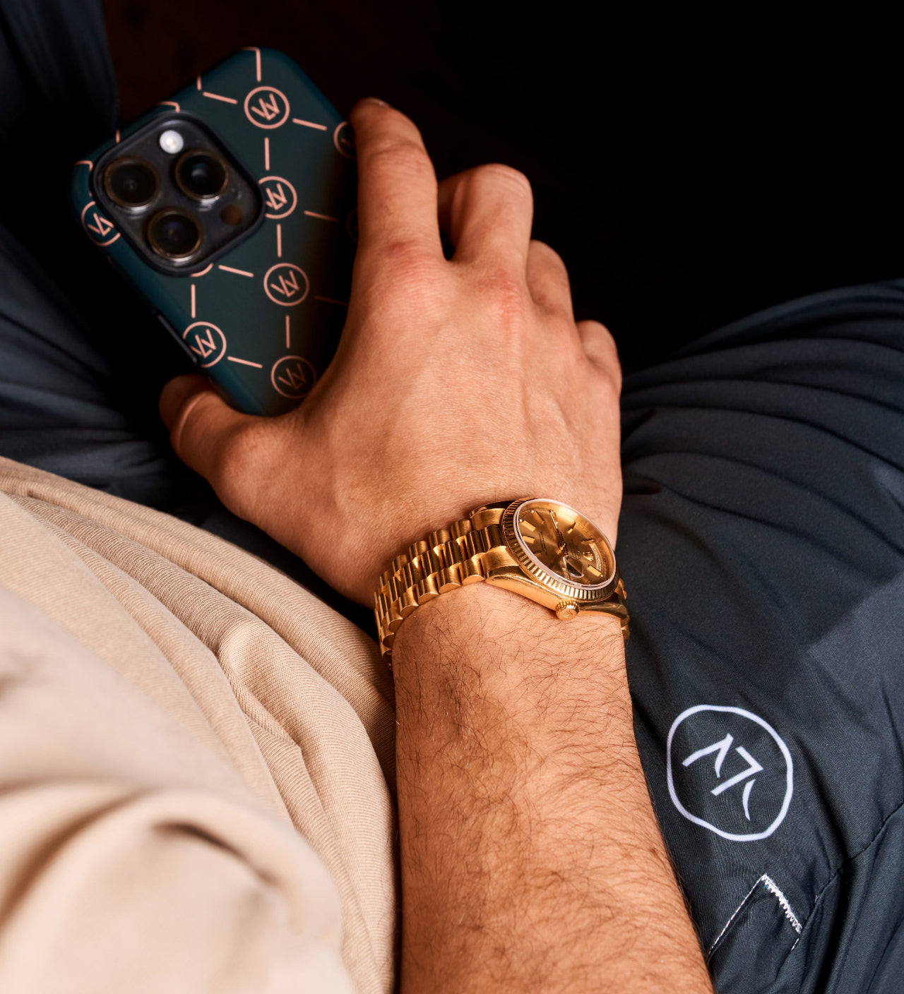 Relaxed lifestyle shot of a person wearing a gold wristwatch and holding a Luxewrist phone case, with branded navy trackpants and beige sweatshirt visible