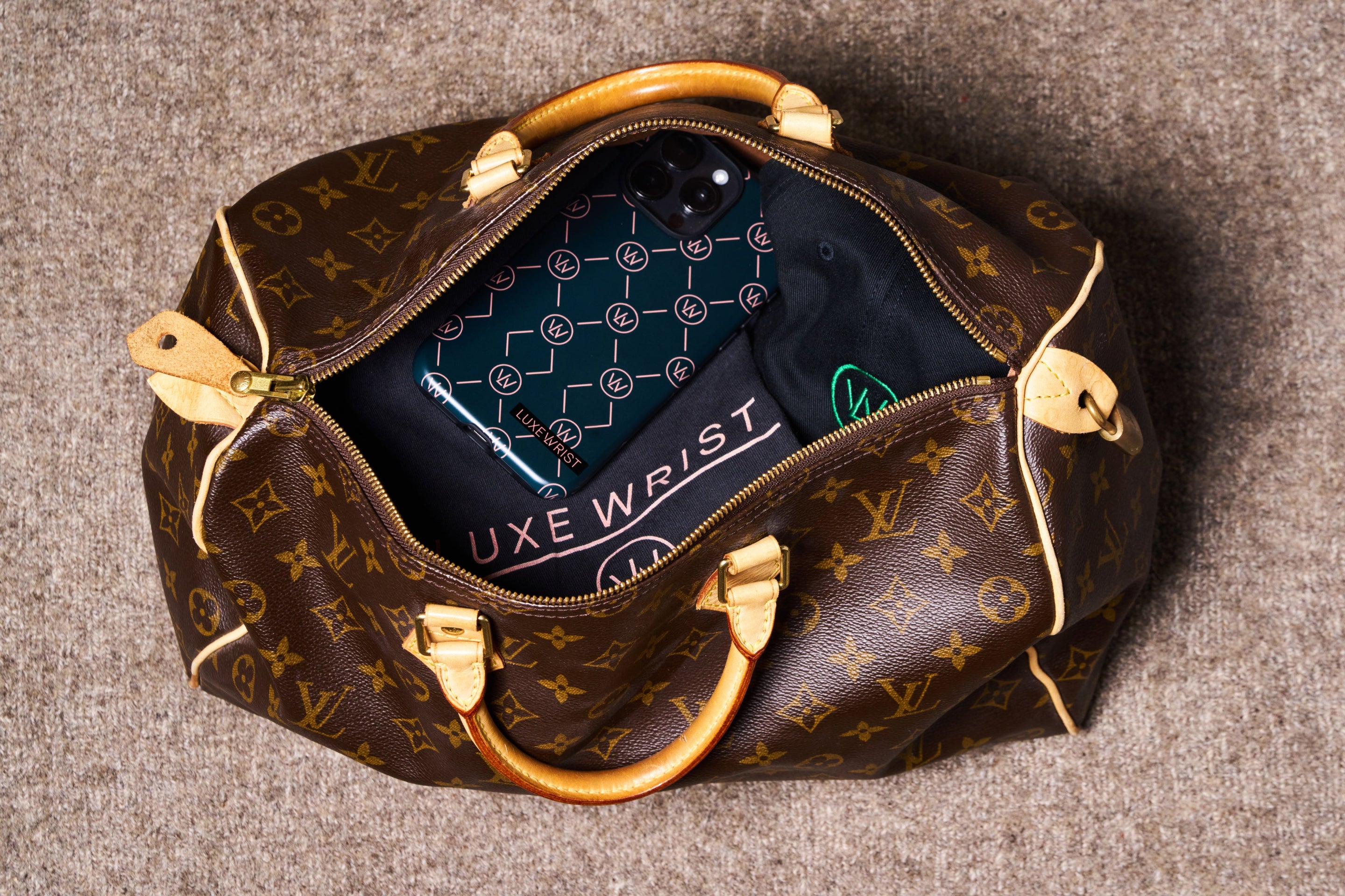 Top-down view of an open designer duffle bag containing a Luxewrist teal logo phone case, neatly folded black t-shirt and cap with Luxewrist logo